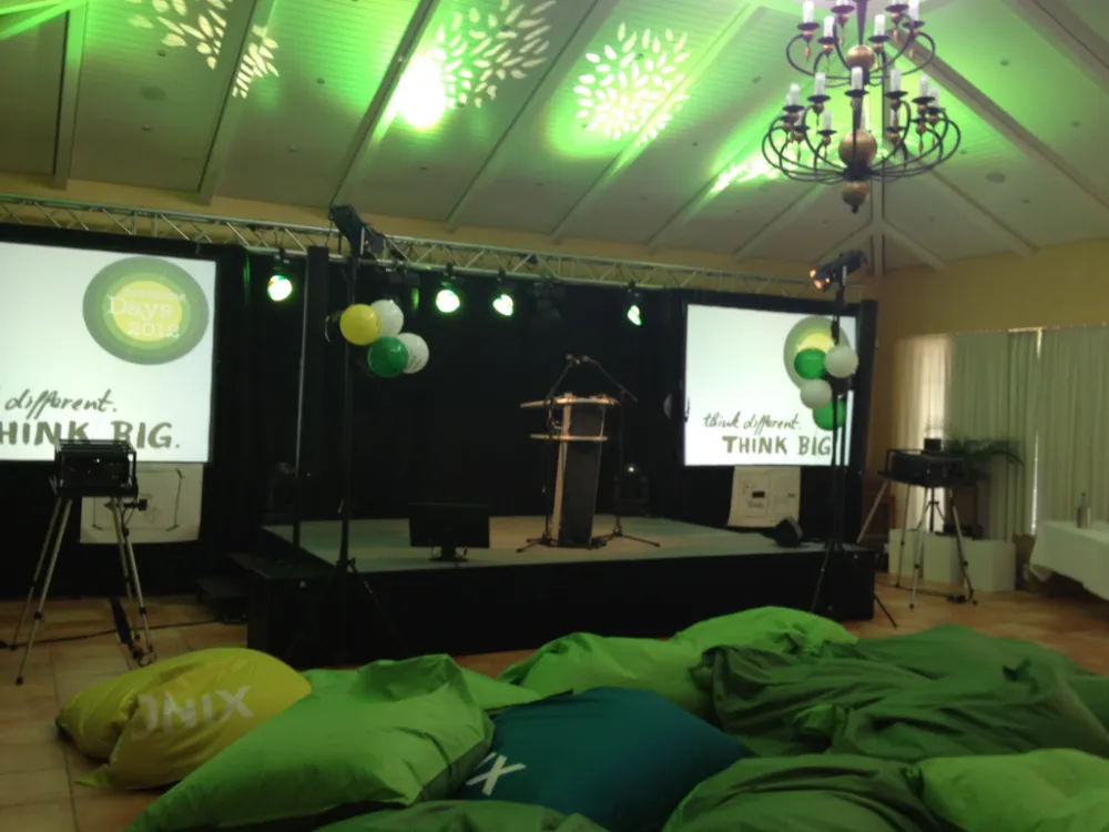 Event stage setup featuring a podium and two screens displaying 'Think Big' and 'Prototyping Days 2012' logos, illuminated by green ceiling lights, with a casual audience area filled with green and yellow beanbag chairs