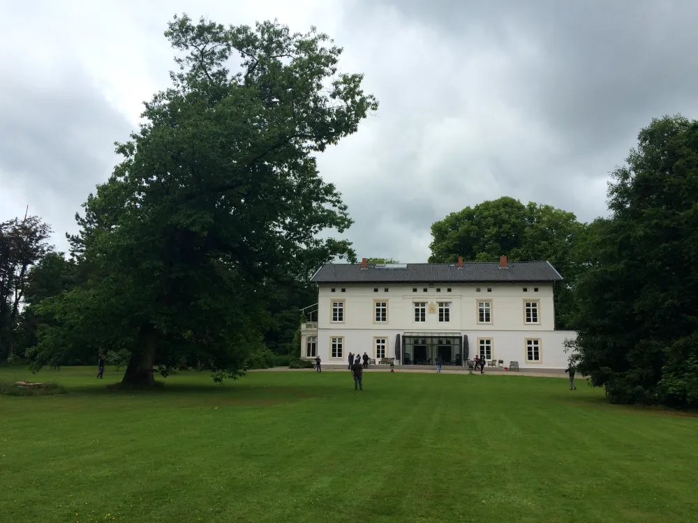 White two-story manor house situated behind a vast green lawn with a large, leaning tree on the left and small groups of people walking near the entrance under a cloudy sky