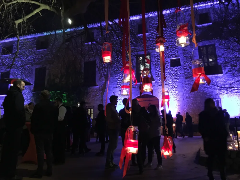 Nighttime outdoor event featuring hanging candle lanterns with red ribbons, a crowd of people socializing, and a stone building illuminated by blue and purple lights in the background