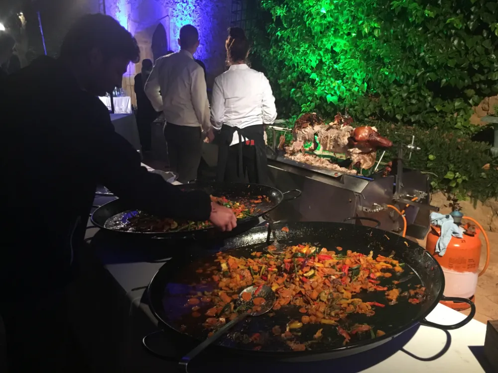 Outdoor catering station at night featuring large paella pans filled with vegetable dishes and a whole roasted pig on a spit, with chefs in white uniforms standing in the background