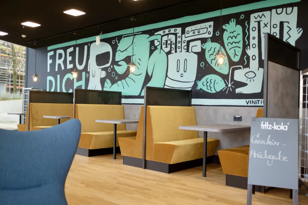 Modern canteen interior featuring mustard-yellow booth seating and a large black and mint-green mural with stylized fish and the text 'FREU DICH', with a dish return sign in the foreground