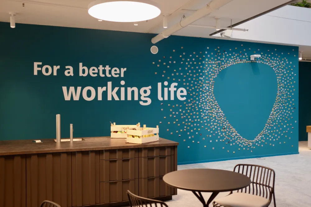 Office break area featuring a teal wall with the text 'For a better working life' and a large mosaic made of small photos, situated behind a kitchen island with fresh fruit and a seating table