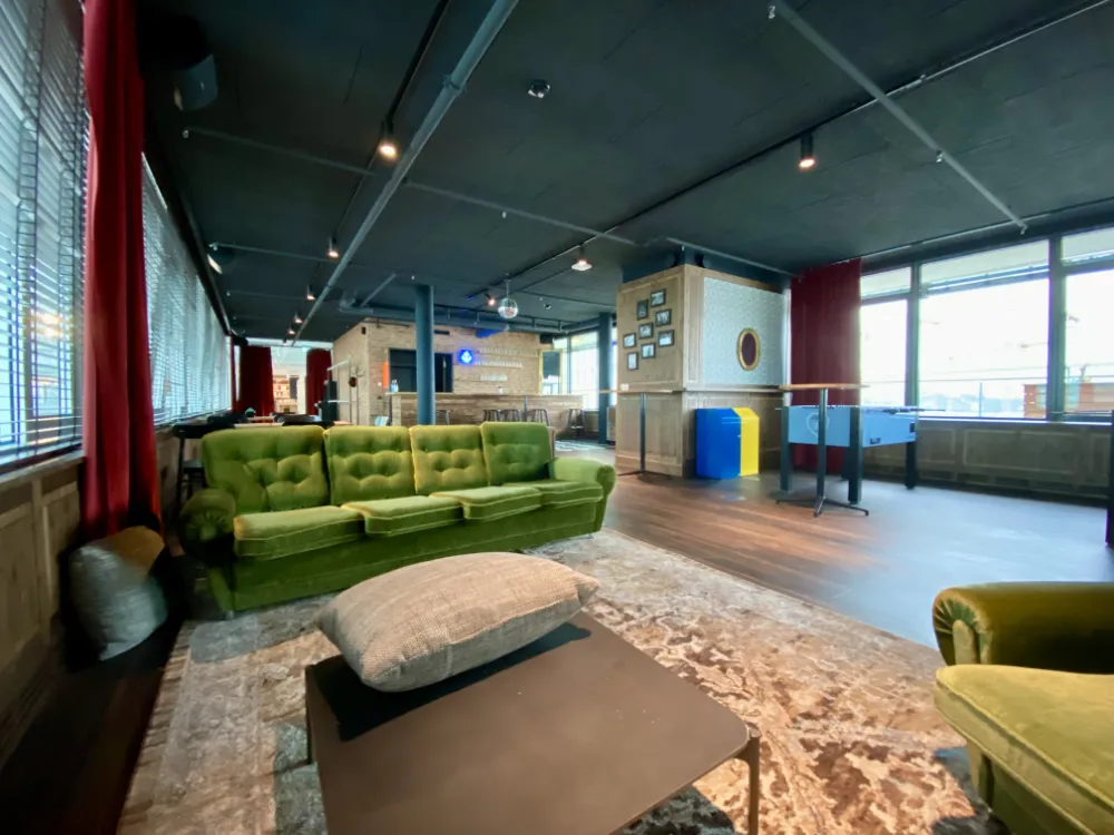 Lounge area in an office with a retro vibe, featuring a green velvet sofa, patterned rug, foosball table, dark ceiling, and a bar counter in the background