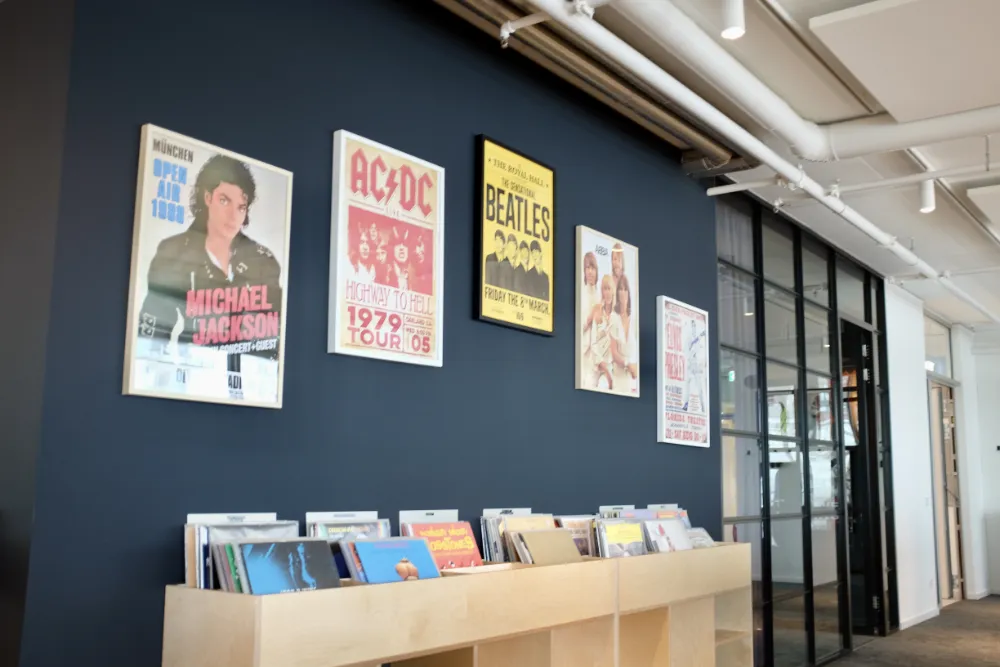 Music-themed corner in a modern office featuring framed concert posters of Michael Jackson, AC/DC, The Beatles, and ABBA mounted on a dark blue wall above a light wooden record display stand