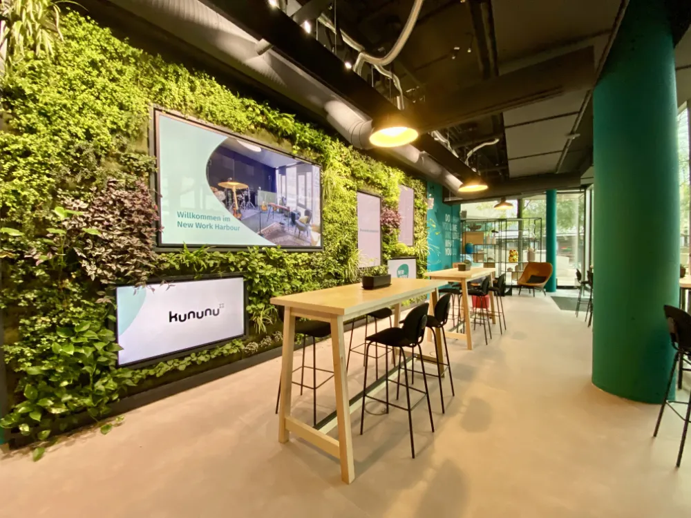 Modern office lobby area featuring a lush vertical garden wall mounted with digital screens displaying 'kununu' and 'Willkommen im New Work Harbour', furnished with high wooden tables and black bar stools